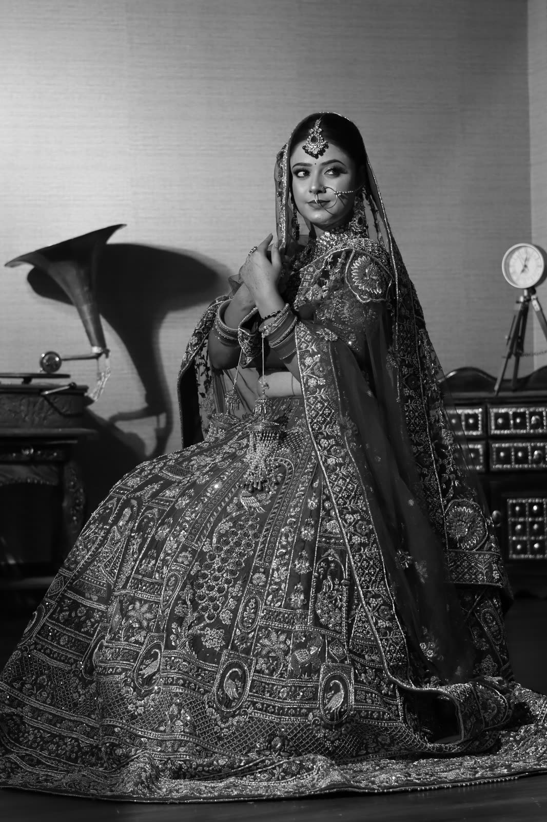 Black and white portrait of a bride in an embroidered lehenga beside a vintage gramophone